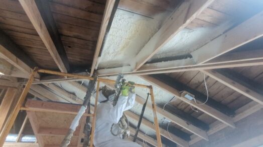 Worker in protective suit sprays foam insulation onto the underside of a wooden roof structure from scaffolding inside a building under construction.