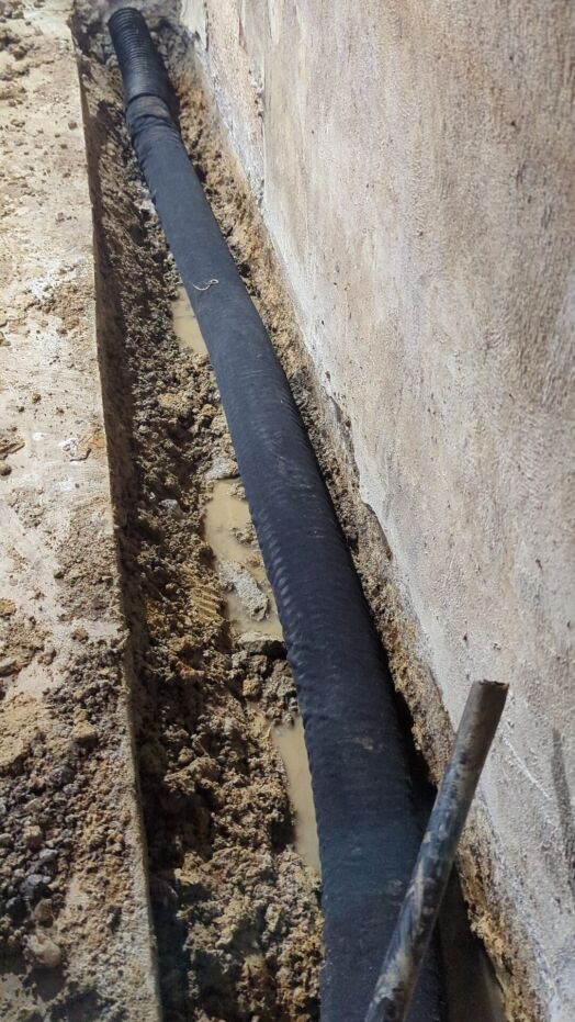 A black drainage pipe is installed in a trench along a concrete wall, surrounded by damp soil and some standing water.