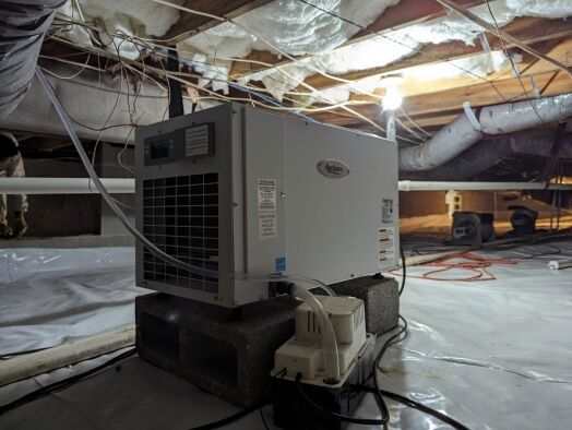 A dehumidifier placed on concrete blocks operates in a crawl space with exposed insulation, ducts, and plastic sheeting covering the ground.