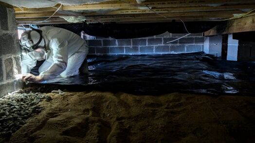 A person in protective gear works in a crawl space with a dirt floor, placing plastic sheeting against the wall.