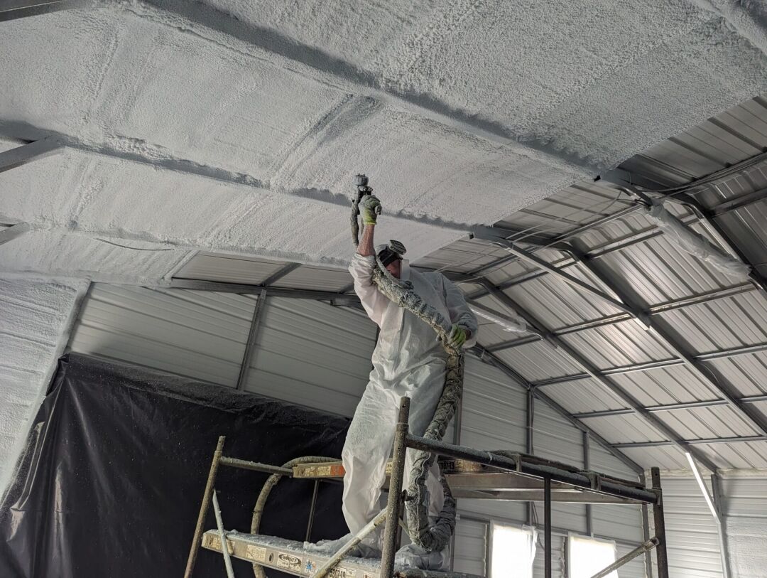 A worker in protective gear sprays insulation foam onto the ceiling of a metal building while standing on scaffolding.