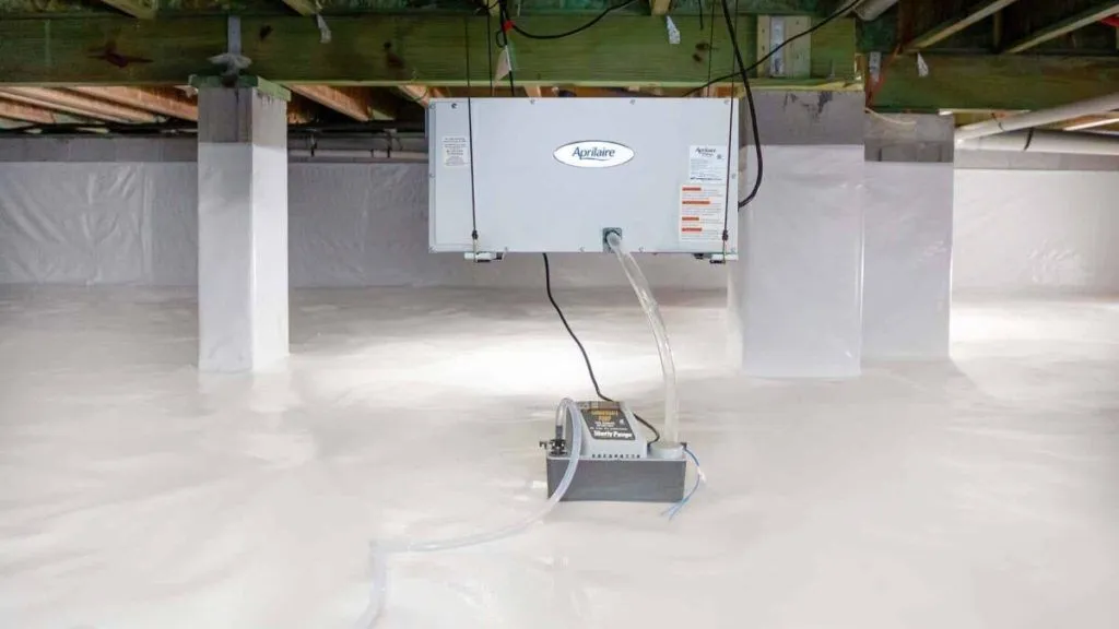 A dehumidifier is installed in a clean, sealed crawl space with white vapor barrier lining the floor and support columns.