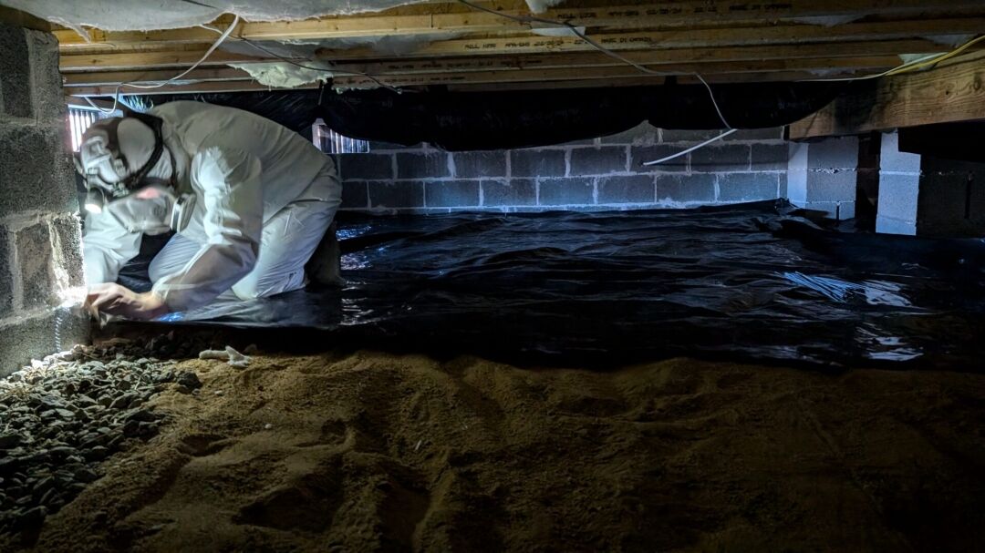 A person in protective gear works inside a crawl space with exposed dirt, rocks, and plastic sheeting along the ground and walls.