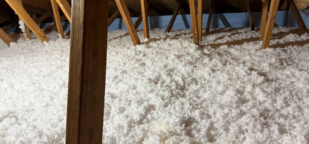 Attic with exposed wooden beams and a floor covered in white blown-in insulation for improved energy savings in Richmond VA.