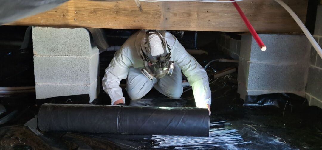 Person in protective suit and respirator installing a plastic vapor barrier under a house crawl space, surrounded by cinder blocks and pipes.