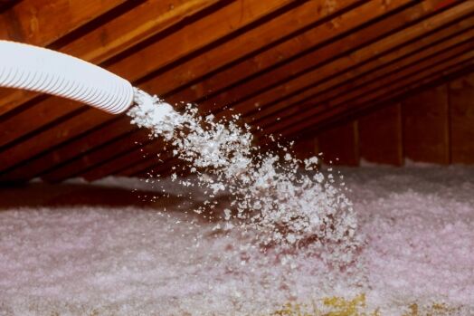 A hose sprays loose-fill insulation in an attic with exposed wooden rafters.