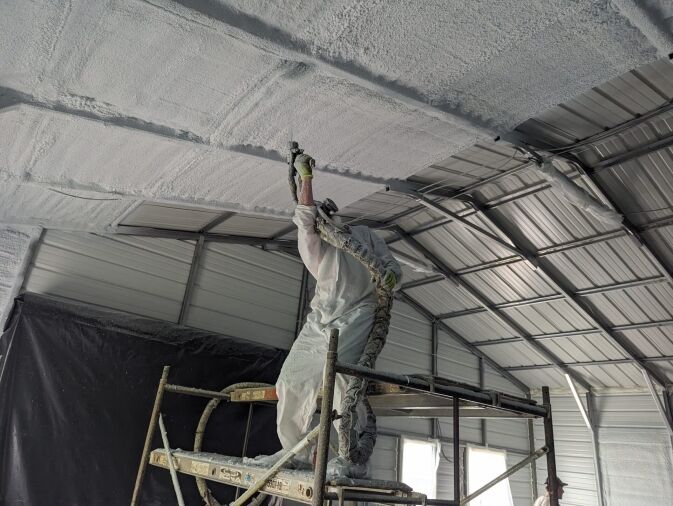 A person in protective gear stands on scaffolding, spraying insulation foam onto the ceiling of a metal building.