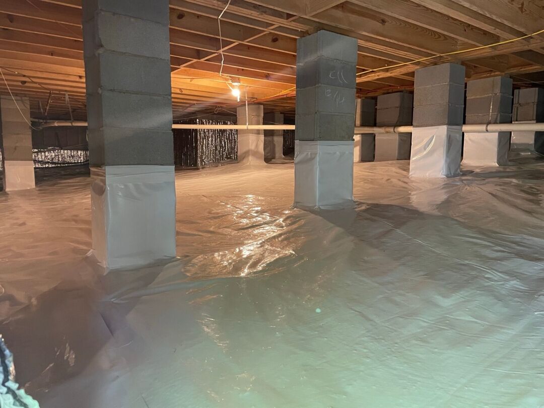 A clean and sealed crawl space with plastic vapor barriers covering the floor and lower walls, supported by concrete block pillars and illuminated by a single hanging light bulb.
