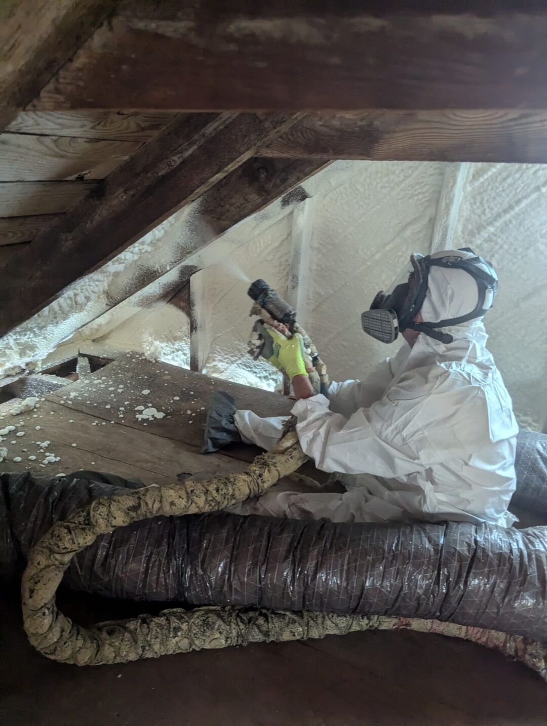 A person in protective gear sprays foam insulation onto the interior roof of an attic space.