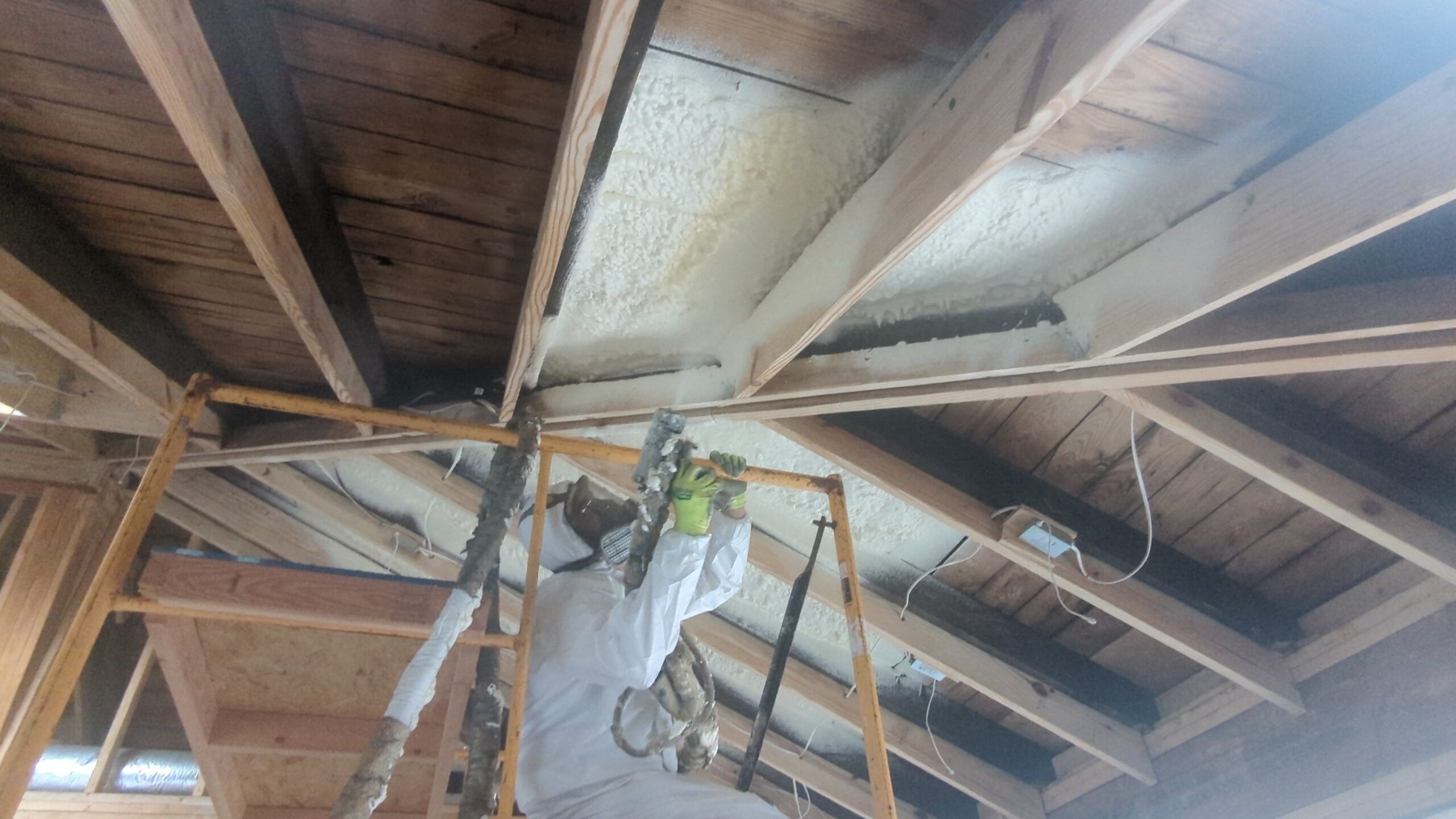 Worker in protective suit sprays foam insulation onto the underside of a wooden roof structure from scaffolding inside a building under construction.