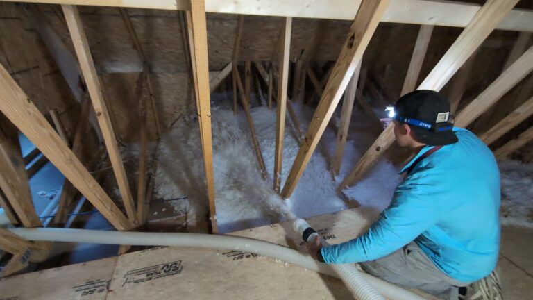 Person in blue shirt uses a hose to spray insulation material between wooden beams in an attic space, wearing a headlamp for visibility.