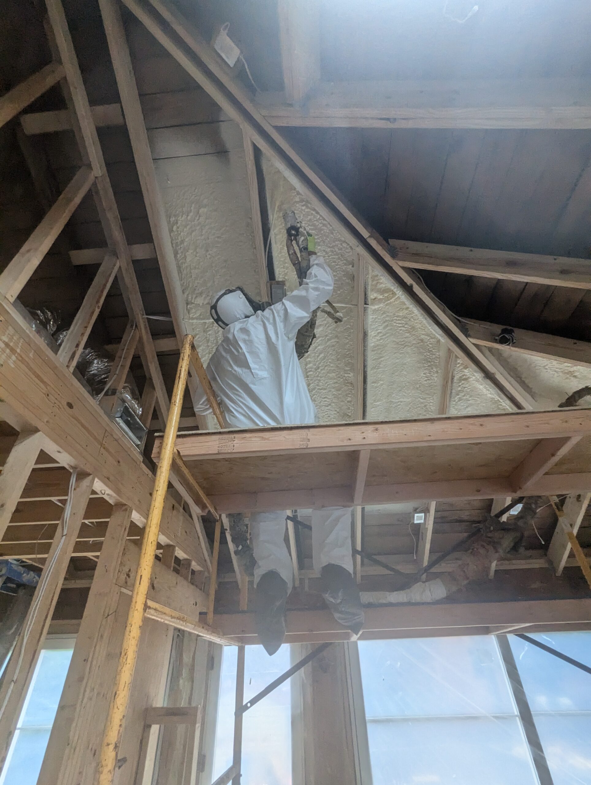 A worker in protective gear sprays foam insulation onto the sloped ceiling of a wooden-framed building under construction.