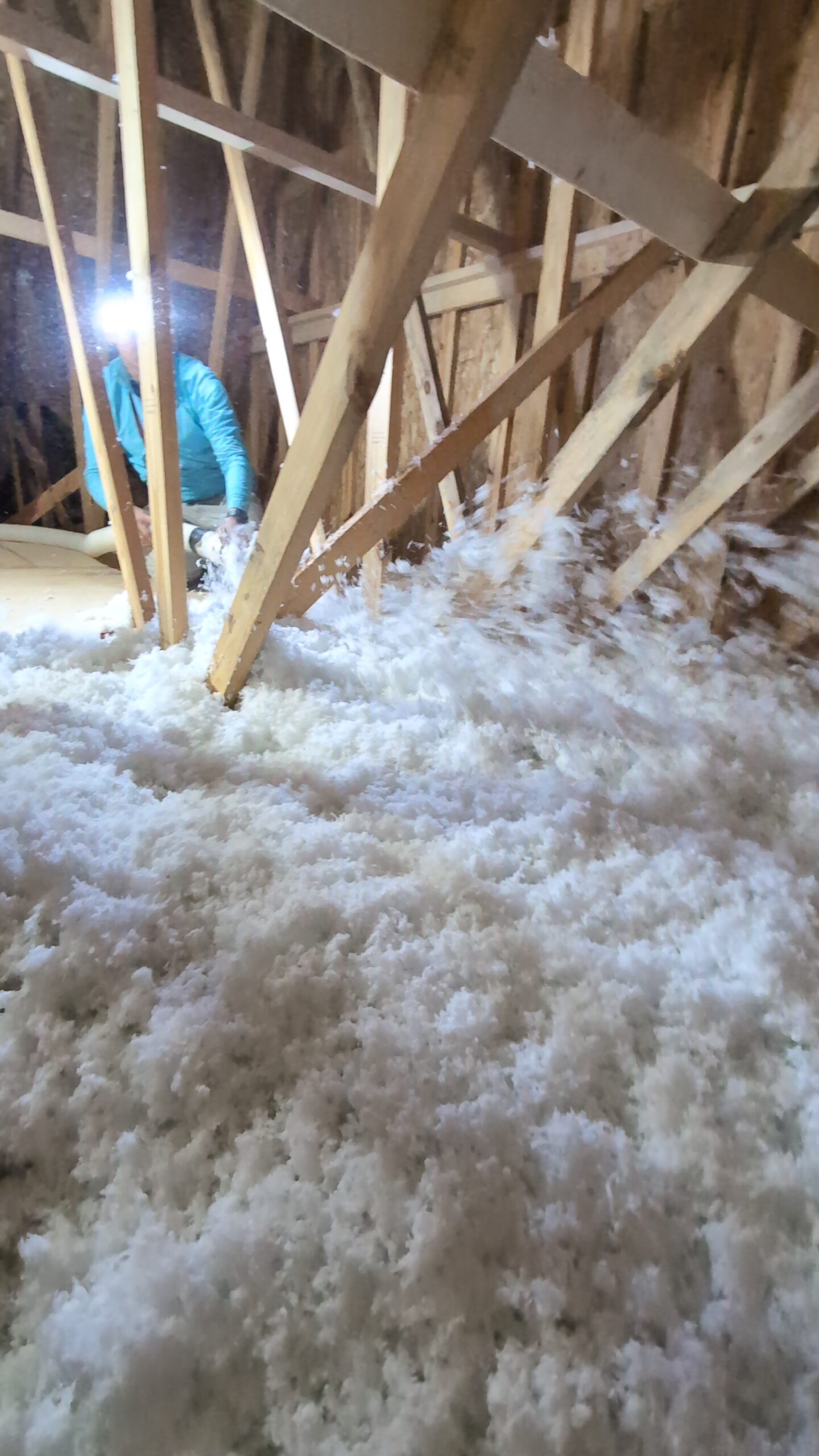 A person with a headlamp is installing or inspecting loose-fill insulation in an unfinished attic with exposed wooden beams.