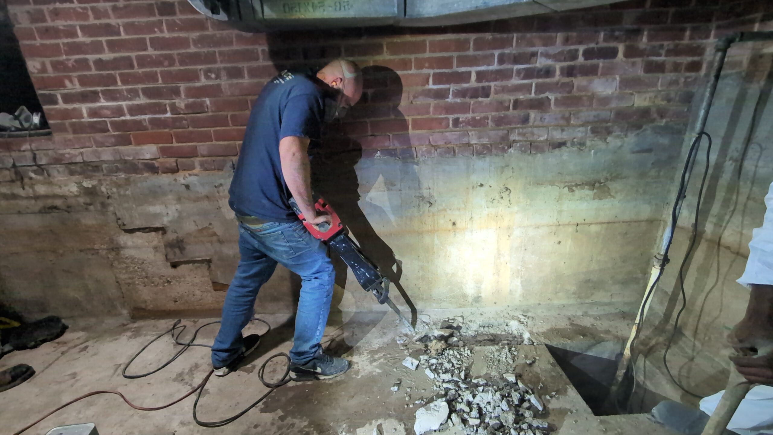 A person uses a power tool to break up concrete on the floor in a basement with exposed brick walls.