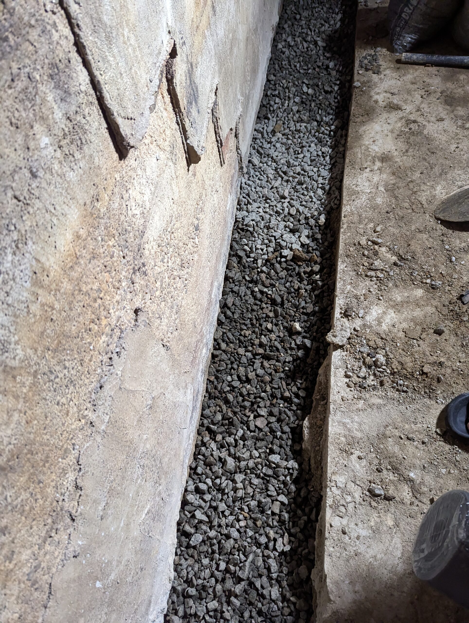 A narrow trench filled with gravel runs alongside a rough concrete wall and floor indoors.