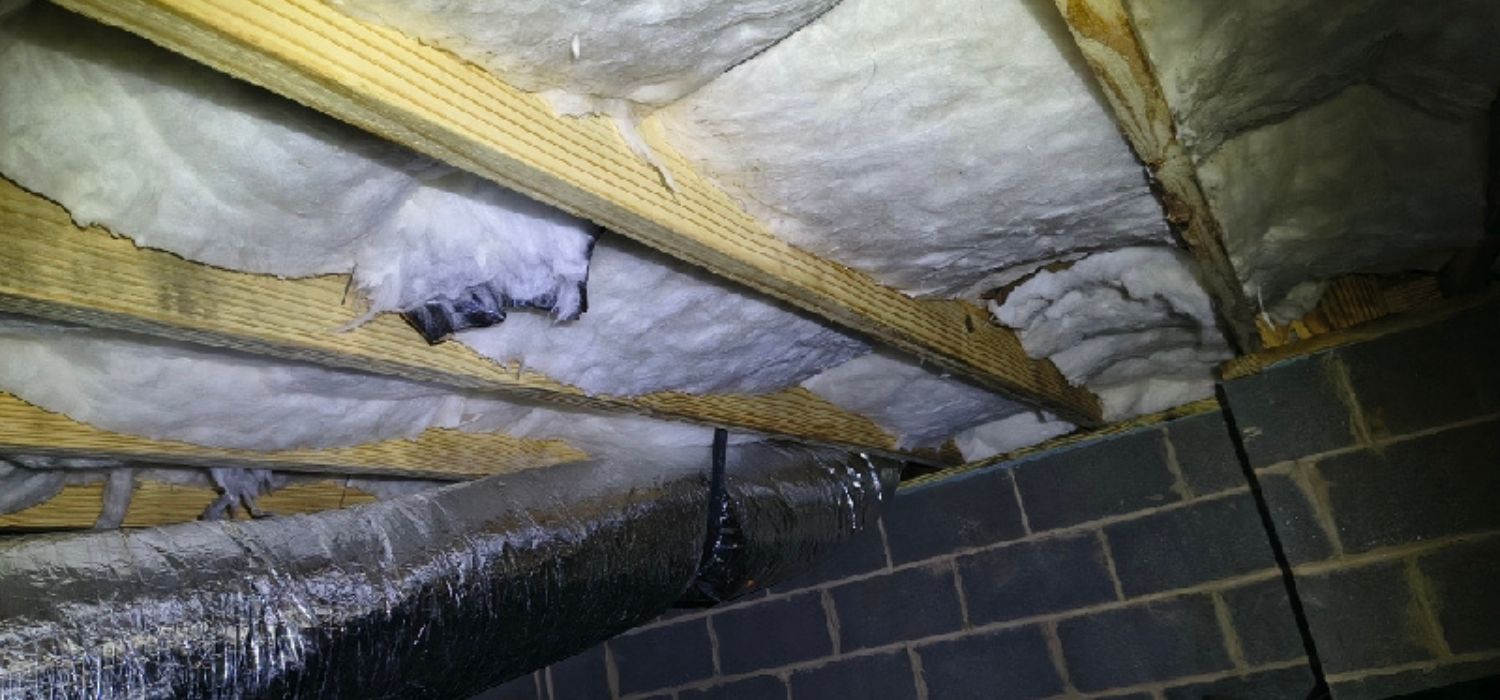 Exposed fiberglass insulation between wooden joists in a basement ceiling next to a metal air duct and unfinished concrete block walls.