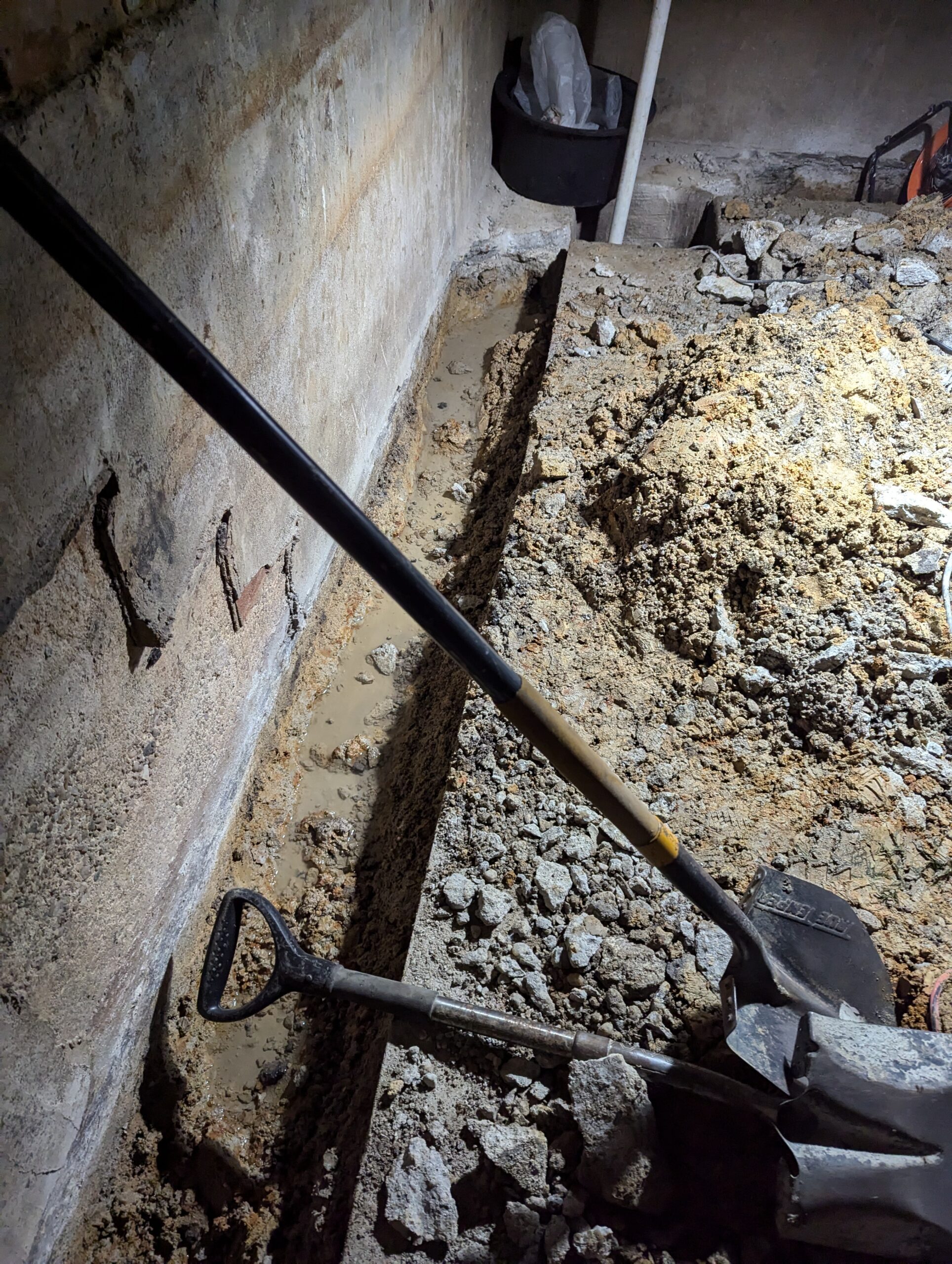 A trench is dug along a concrete basement wall, with a shovel and spade resting on loose dirt and broken concrete.
