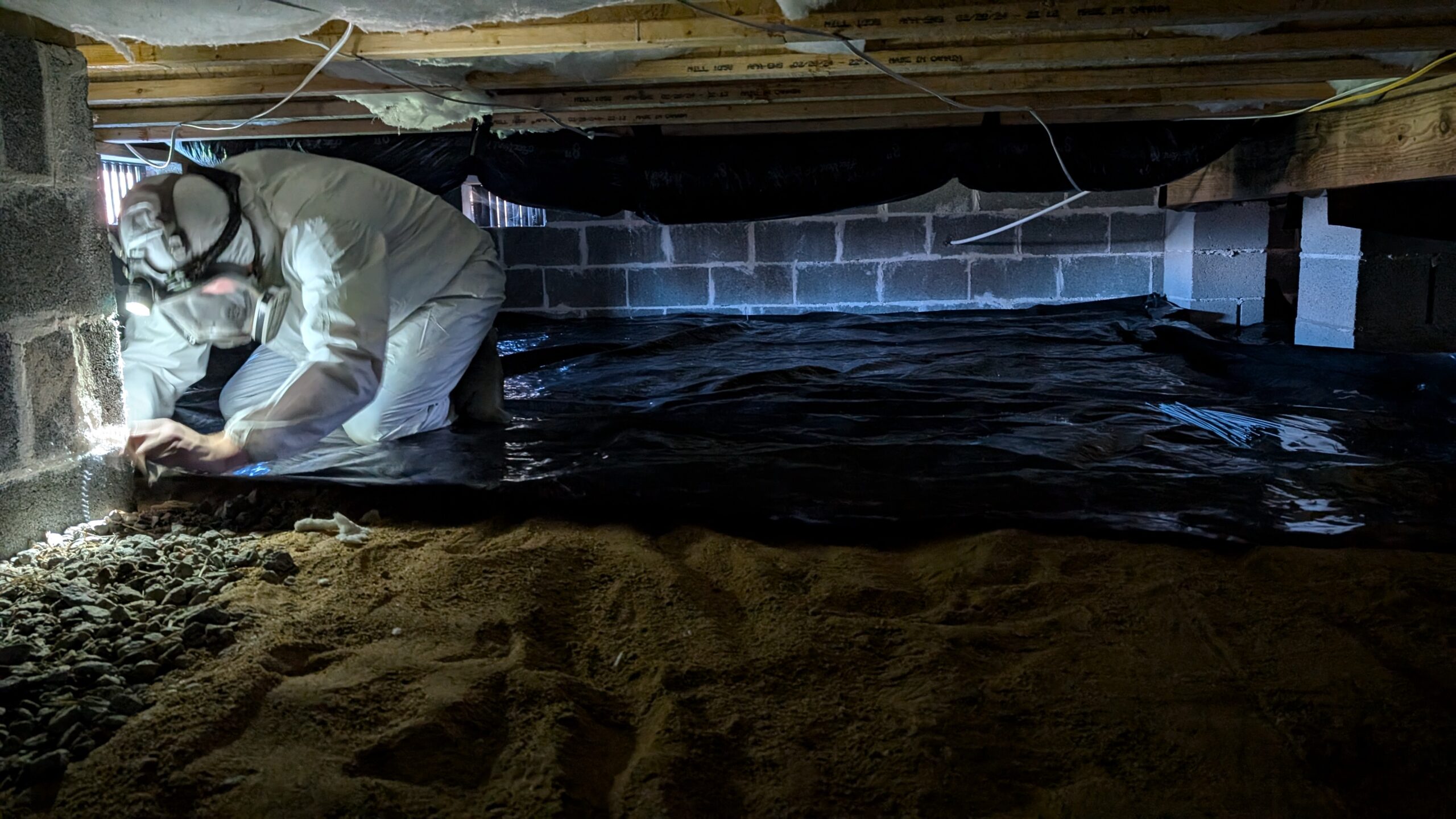 A person in protective gear works in a crawl space with a dirt floor, placing plastic sheeting against the wall.
