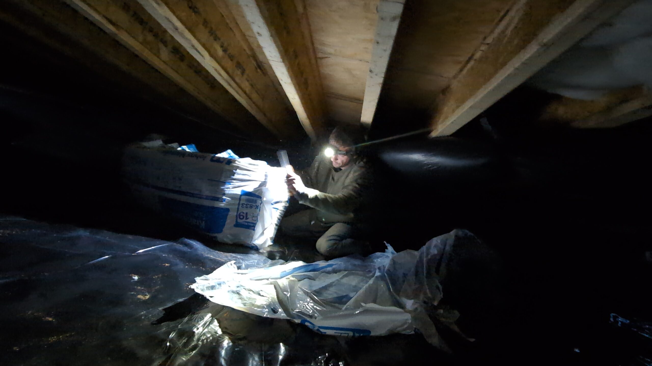Person wearing a headlamp installs insulation in a dimly lit crawl space beneath wooden floor joists, surrounded by plastic sheeting and insulation materials.