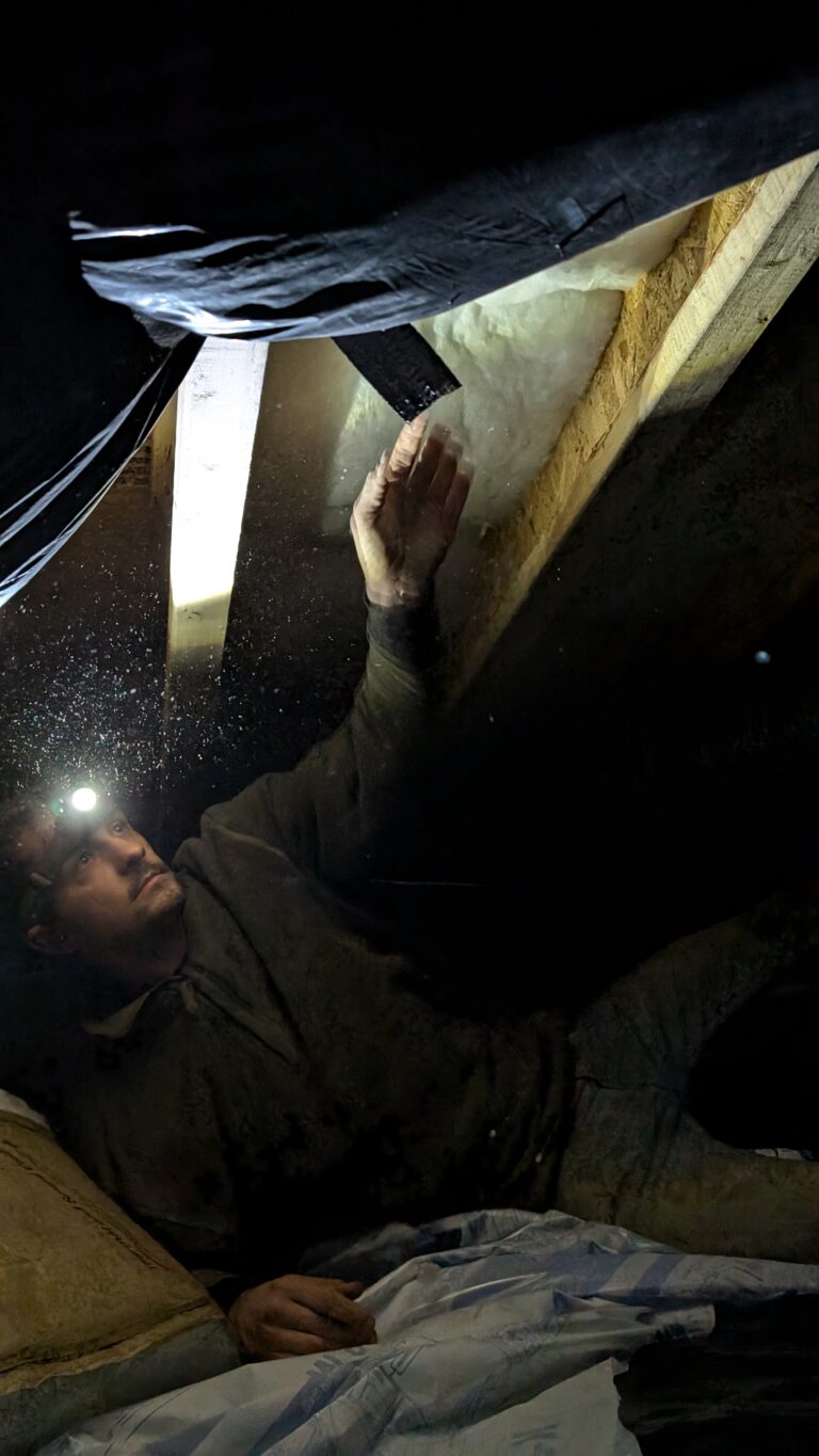 A person wearing a headlamp works on insulation between wooden beams in a dimly lit crawl space.