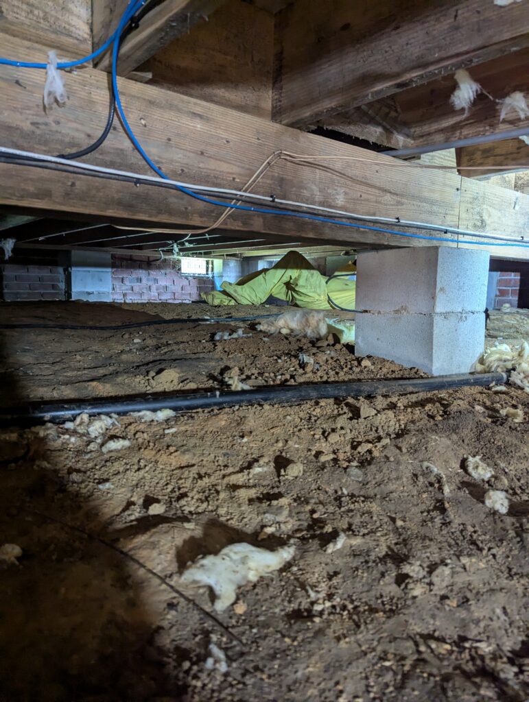 A crawl space with exposed dirt floor, insulation hanging from the ceiling, support beams, blue wiring, and a concrete block pillar.