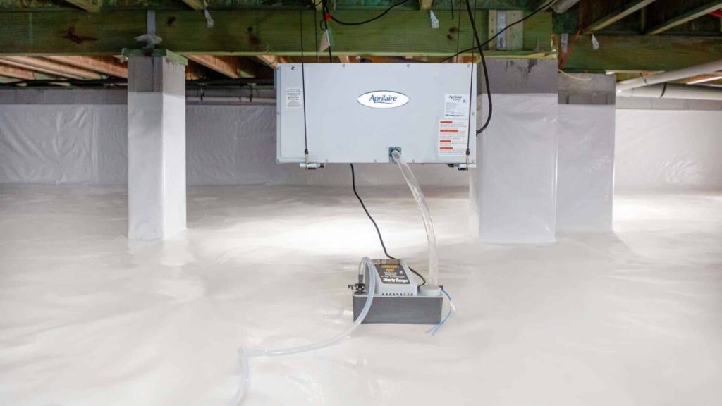 A dehumidifier is installed in a clean, encapsulated crawl space with white vapor barrier lining the floor and support columns.
