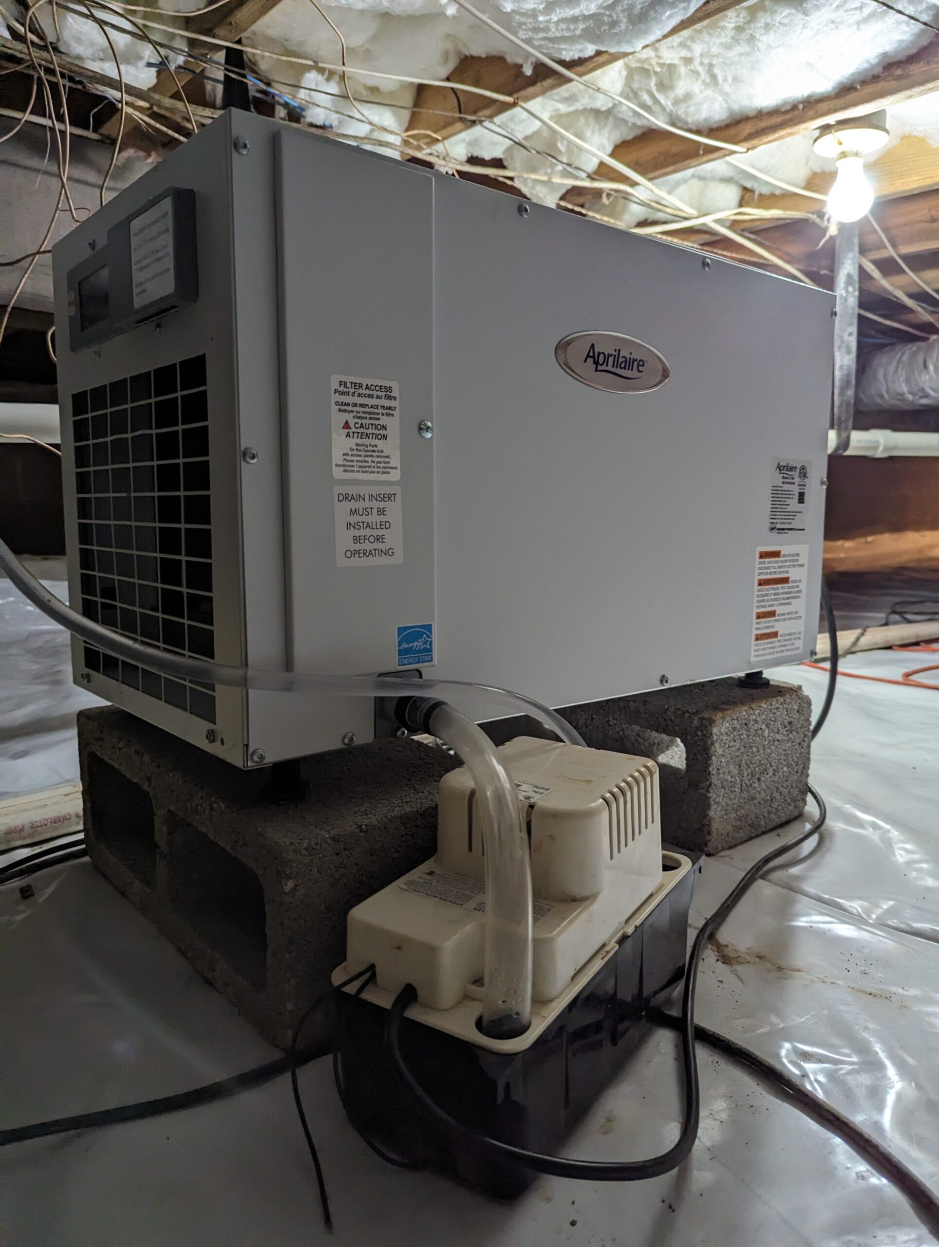 Aprilaire dehumidifier placed on cinder blocks in a crawl space, with attached pump and drainage tubes, surrounded by insulation and exposed beams.