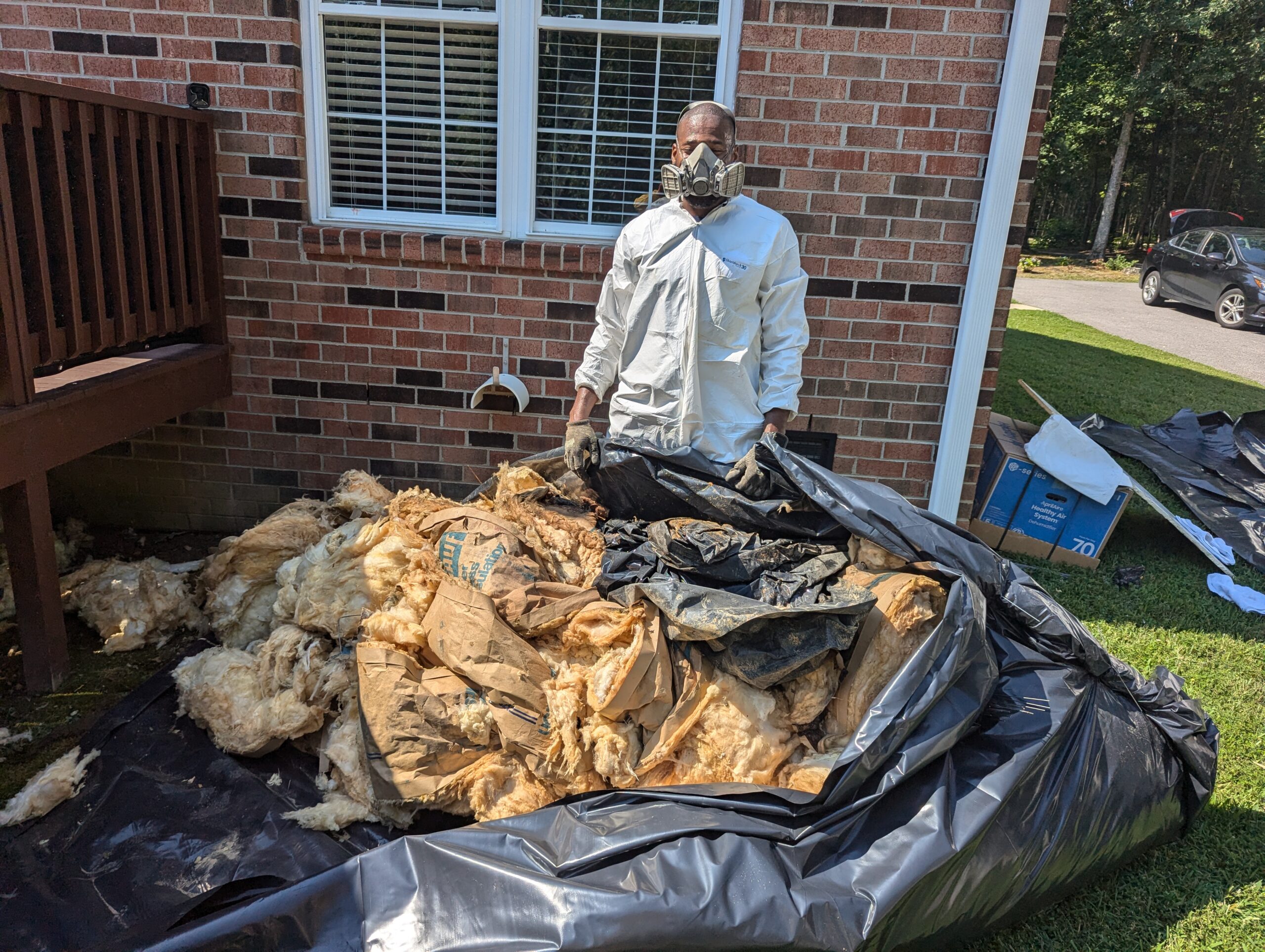 A person in protective clothing and mask stands outside by a pile of removed insulation and debris on a plastic tarp next to a brick house.