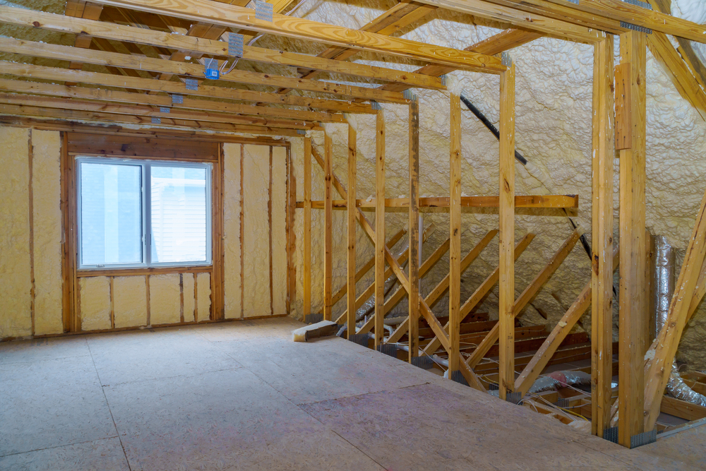 Unfinished attic space with exposed wooden framing, spray foam insulation on the walls and roof, and a window letting in natural light.