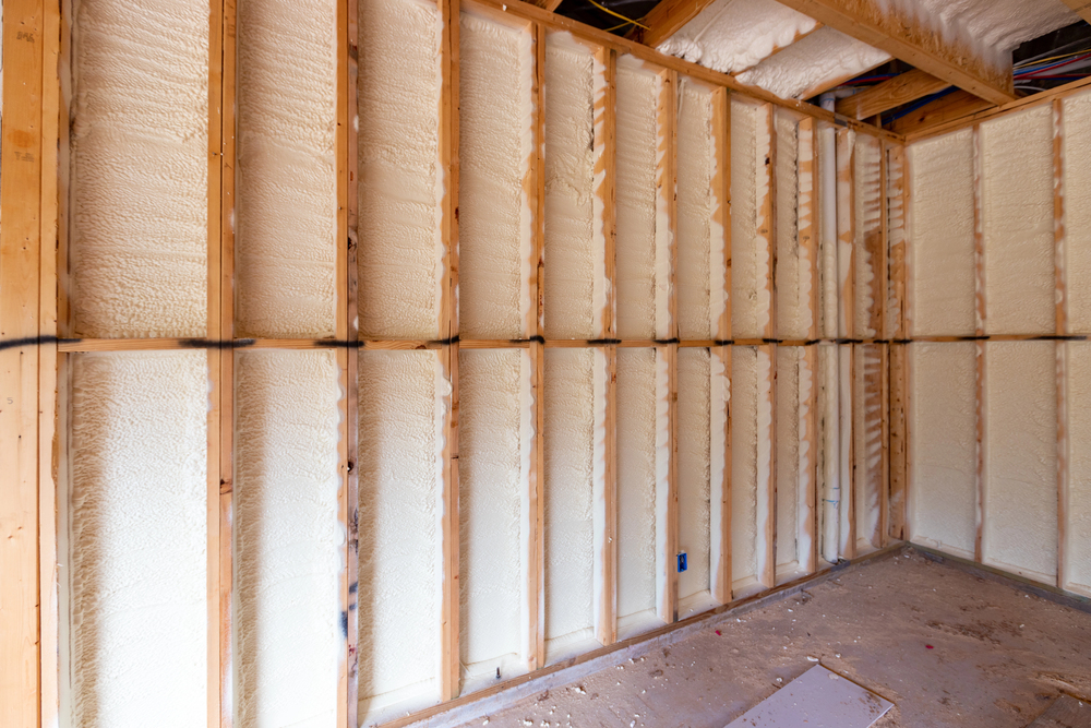 Unfinished room with exposed wooden wall studs and spray foam insulation applied between the studs, with a bare floor and some debris.
