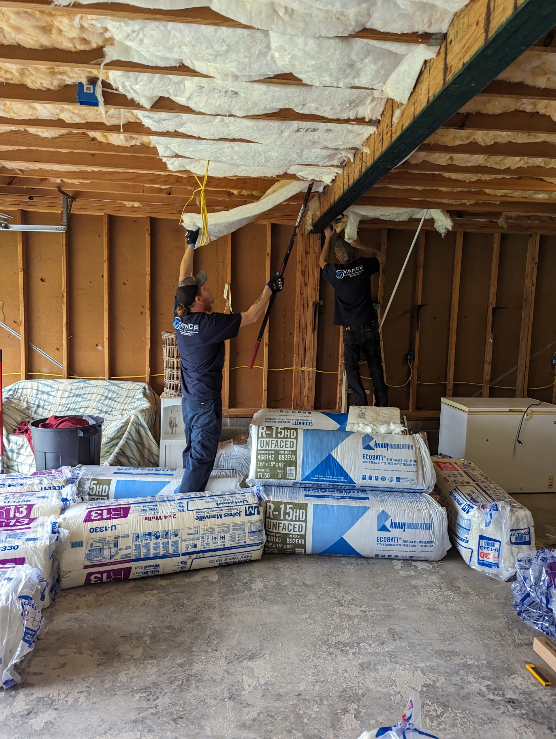 Two workers install insulation in the ceiling of a partially finished room. Bundles of insulation material are stacked on the floor nearby.