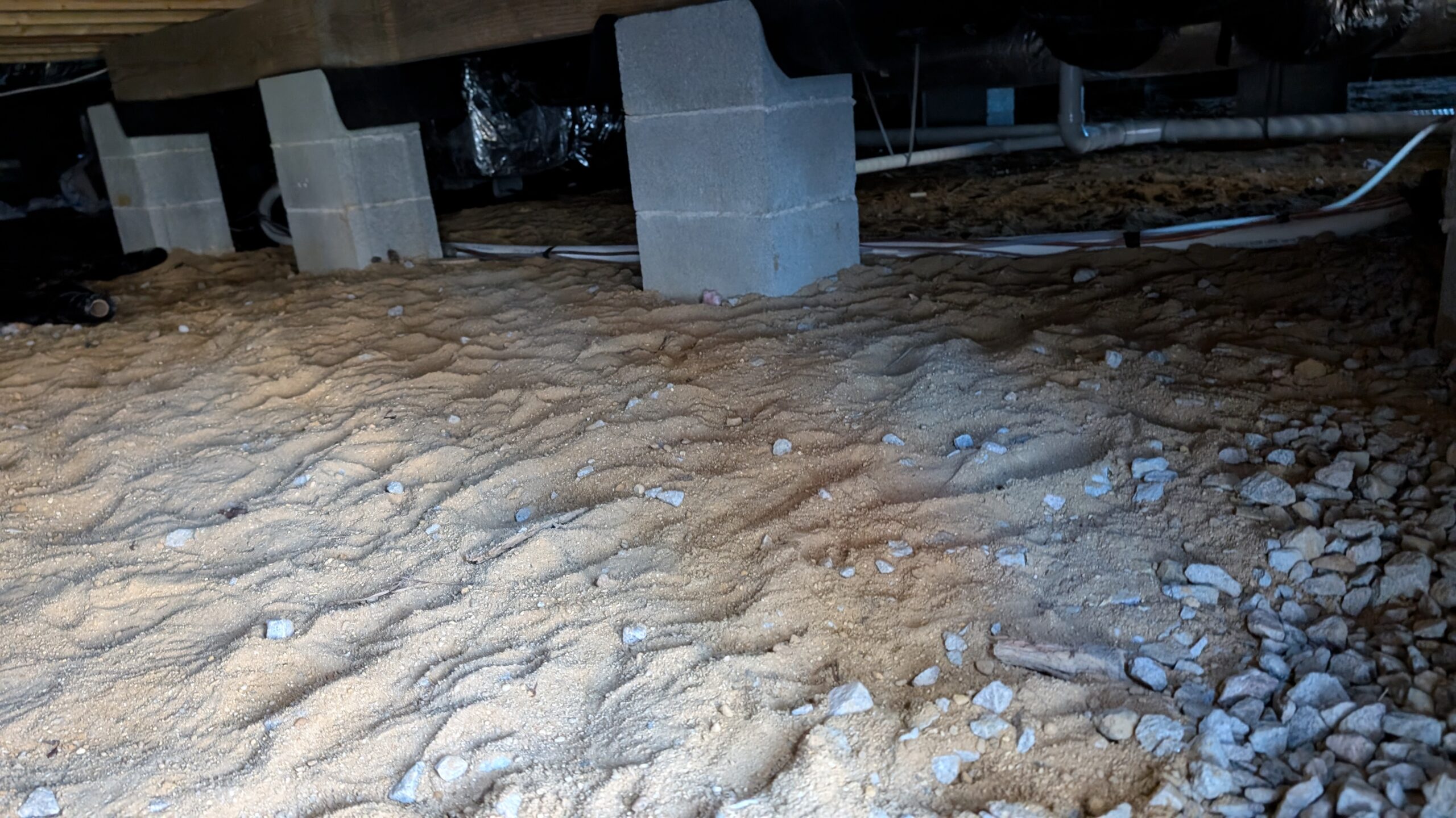 Crawl space under a building with sandy ground, concrete block supports, exposed pipes, and scattered small rocks.