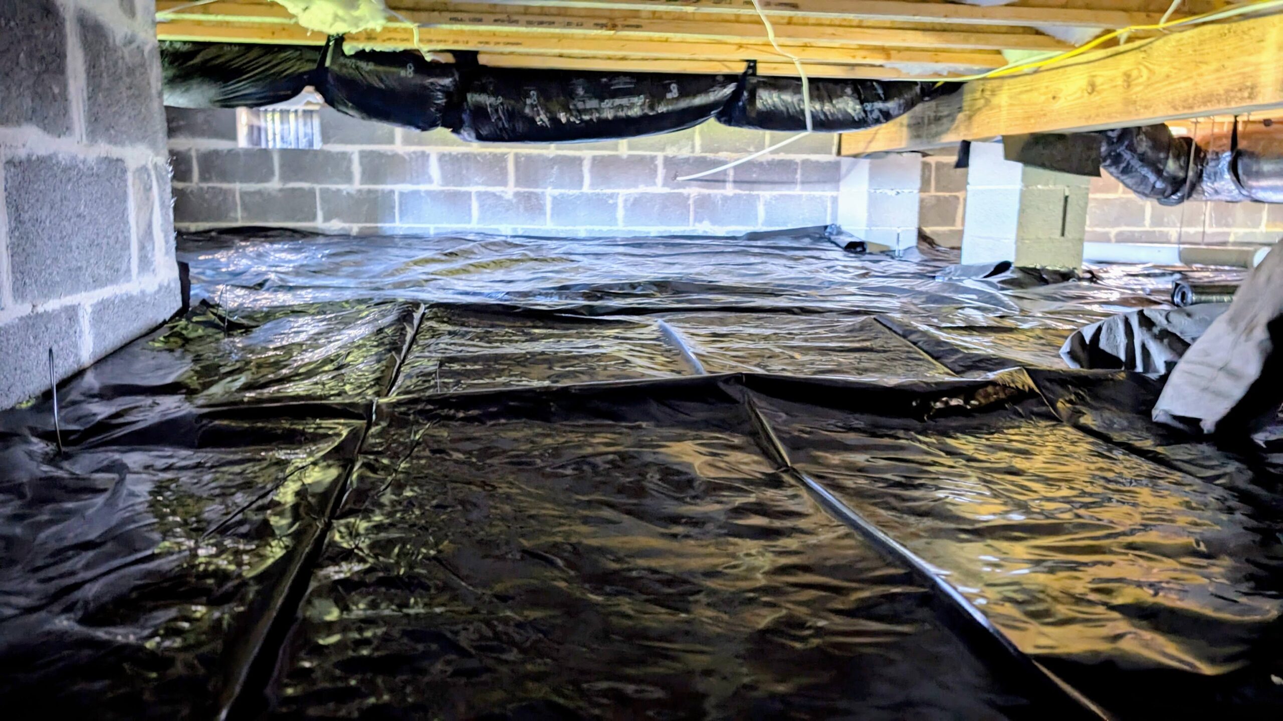 A crawl space with a plastic vapor barrier covering the ground, support beams overhead, and concrete block walls visible along the sides.