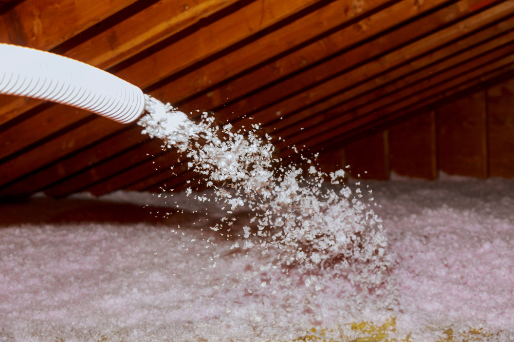 A hose sprays loose-fill insulation in an attic with exposed wooden rafters.
