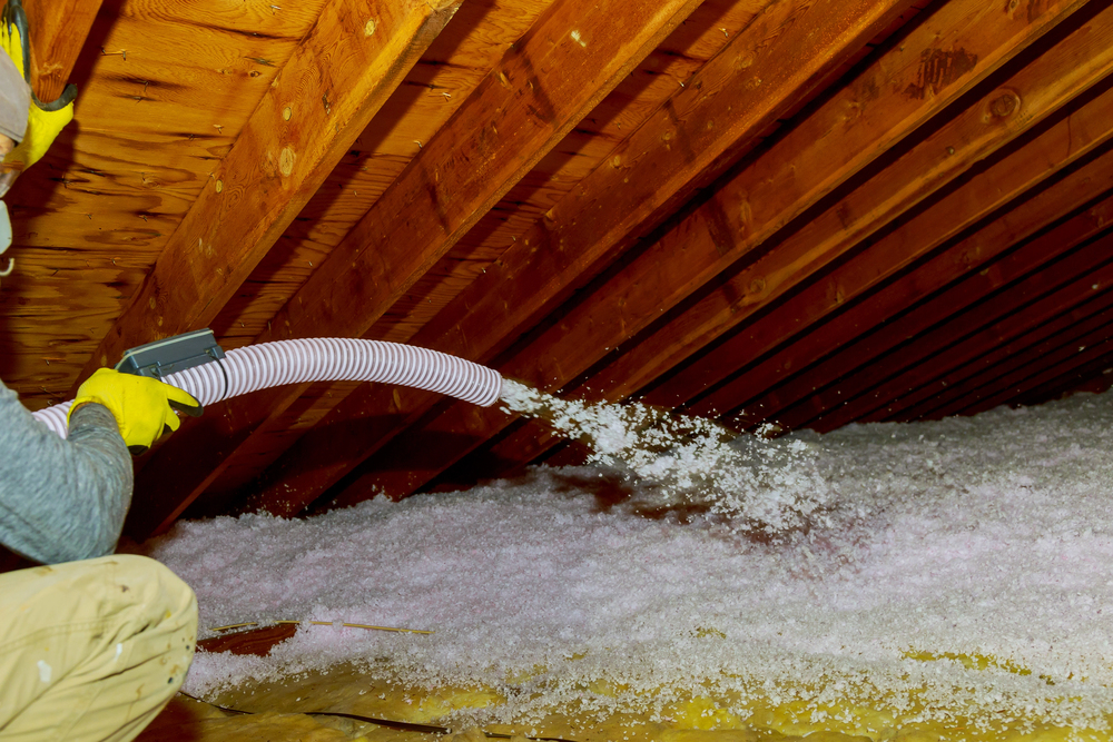A person uses a hose to spray loose fill insulation into an attic, covering the floor between wooden roof beams.