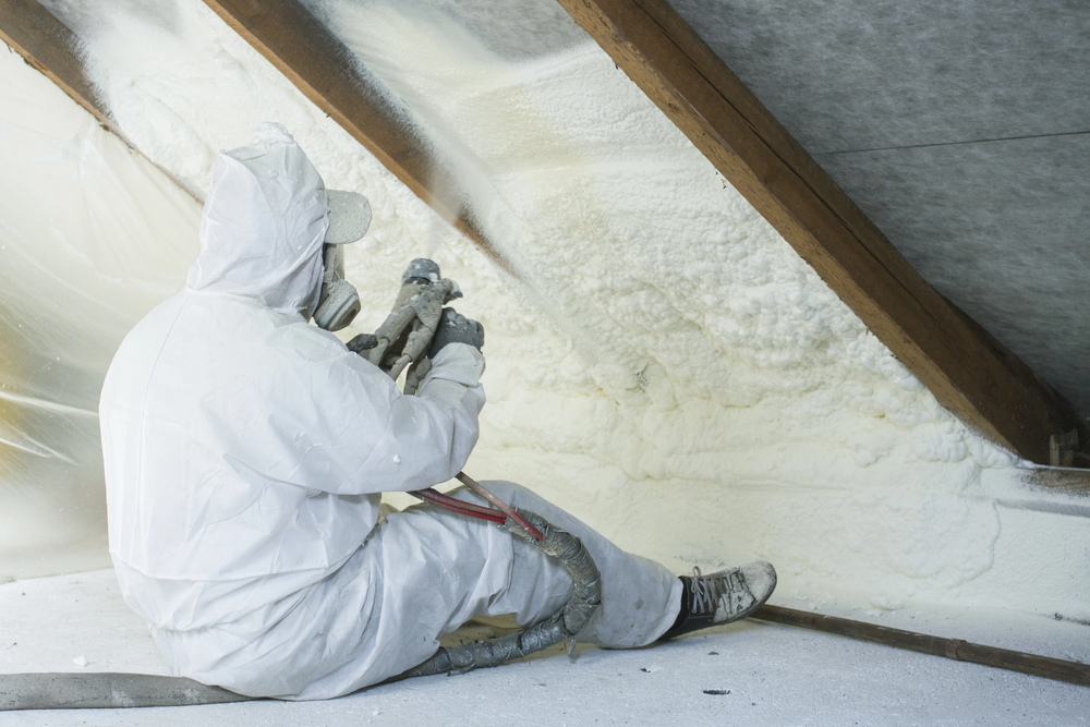 A person in protective gear sprays foam insulation onto the sloped walls of an attic.