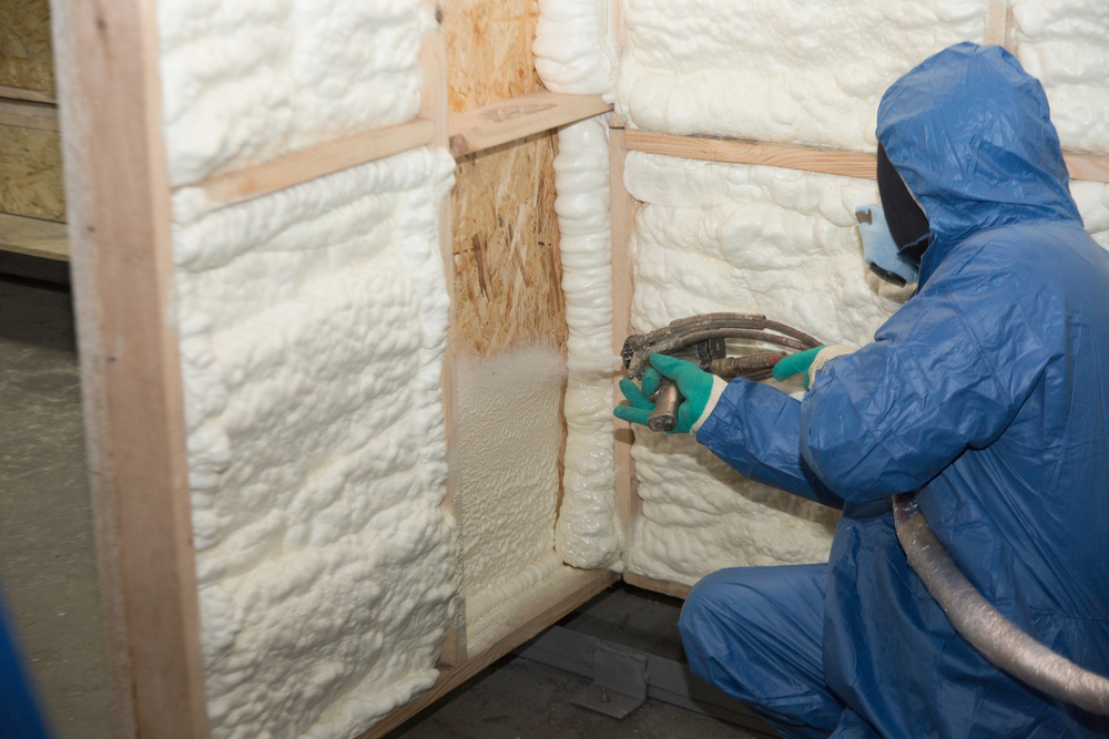 A worker in protective gear sprays foam insulation onto wooden wall panels inside a building.