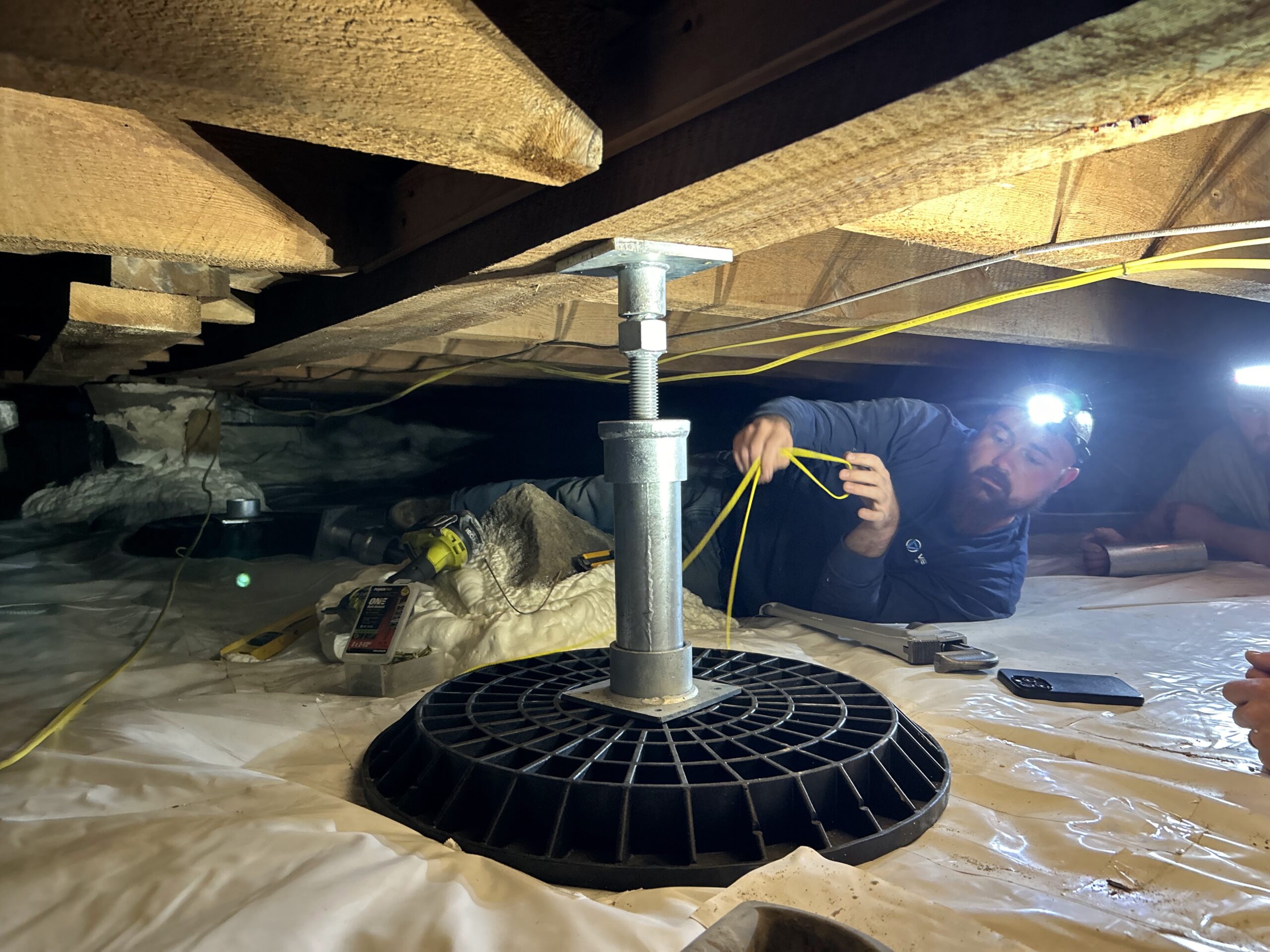 Person wearing a headlamp works under a house crawl space, measuring near a metal support jack on a plastic-covered ground.