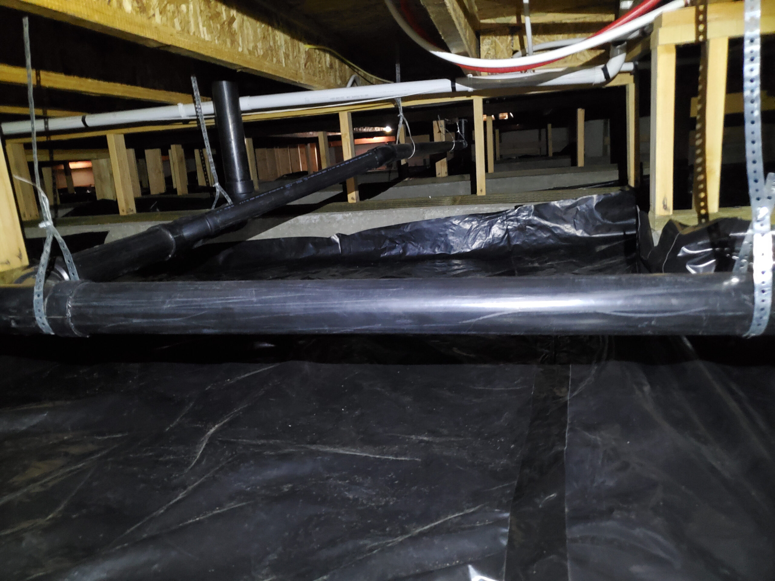 A crawl space with a plastic vapor barrier on the ground, exposed wooden floor joists above, and plumbing pipes secured with metal straps.