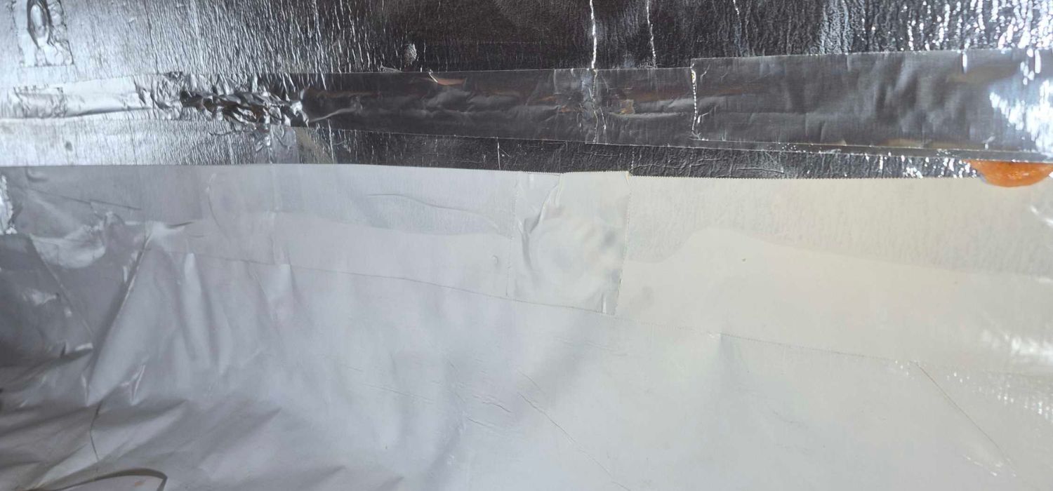 Metallic and white sheets of tape are applied to a flat surface, possibly insulation or a wall, with some orange material visible at the edge.