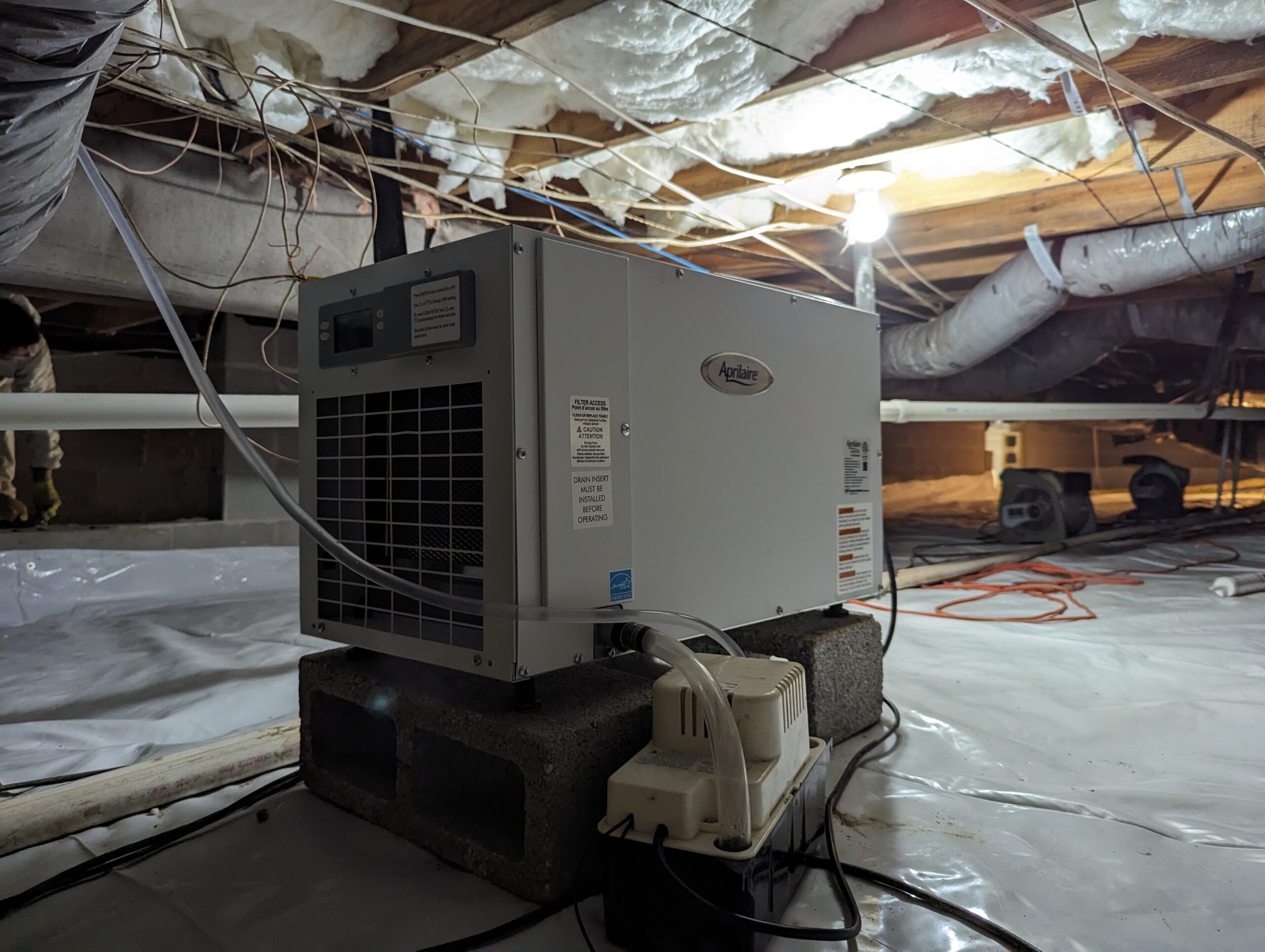 A dehumidifier placed on concrete blocks operates in a crawl space with exposed insulation, ducts, and plastic sheeting covering the ground.