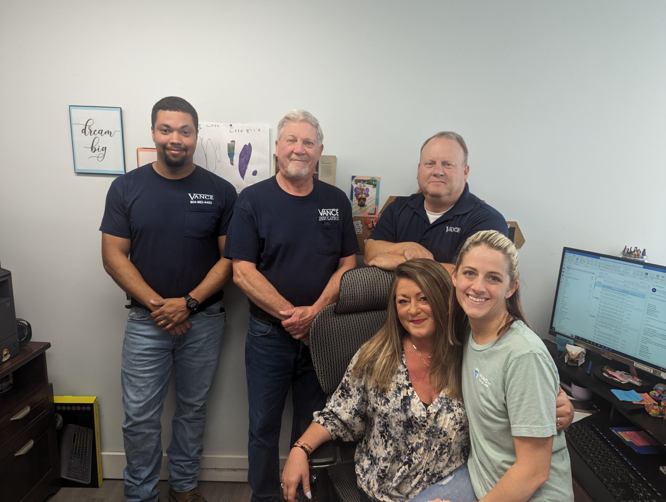 Five people, three men standing and two women sitting, pose for a group photo in an office with computers, artwork, and motivational wall decor visible.