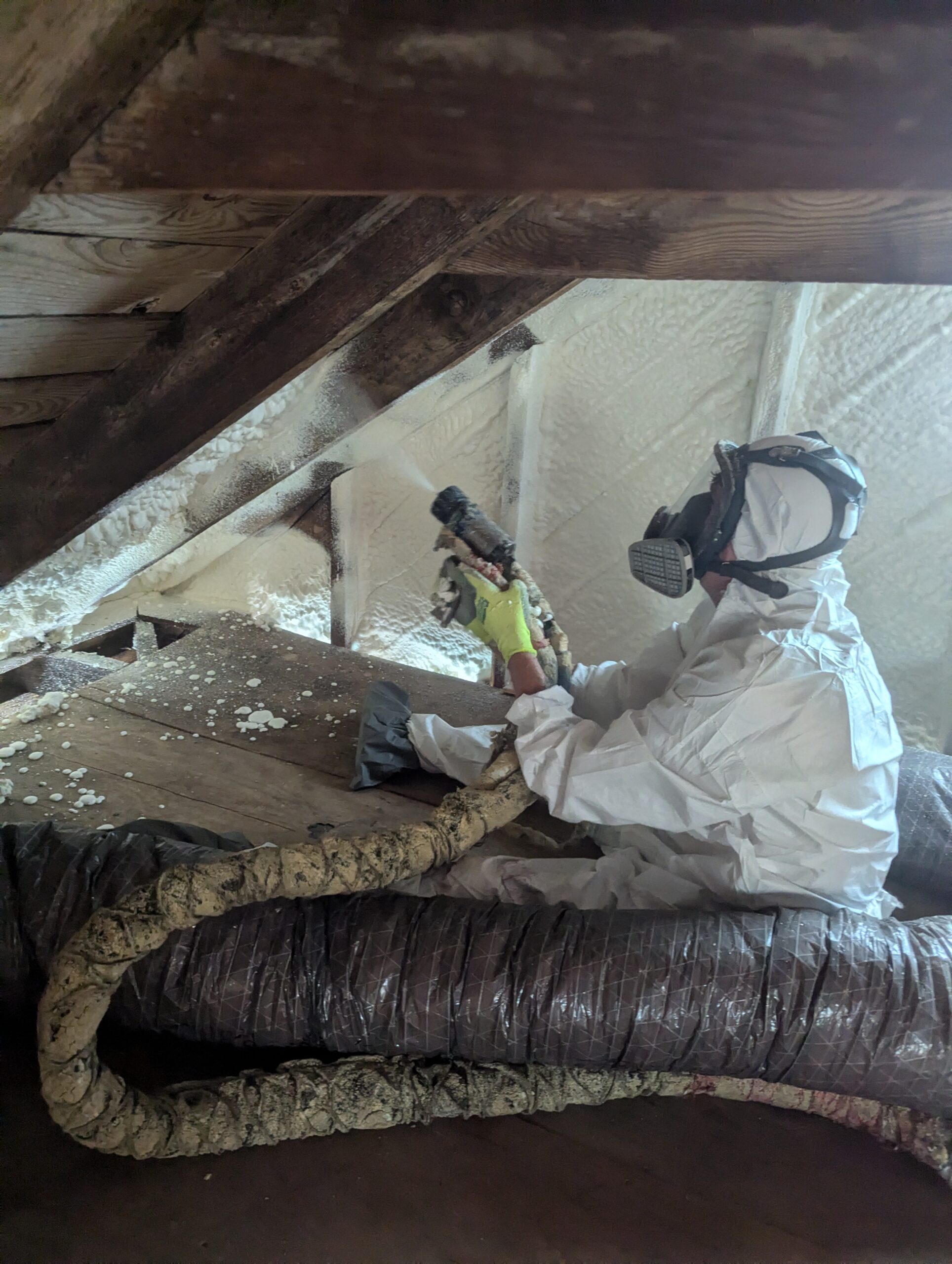 A person in protective gear sprays foam insulation onto the interior roof of an attic space.